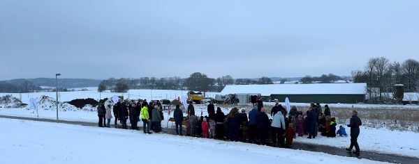 En stor gruppe mennesker samlet til første spadestik i snelandskab med gravemaskine og byggeplads i baggrunden.